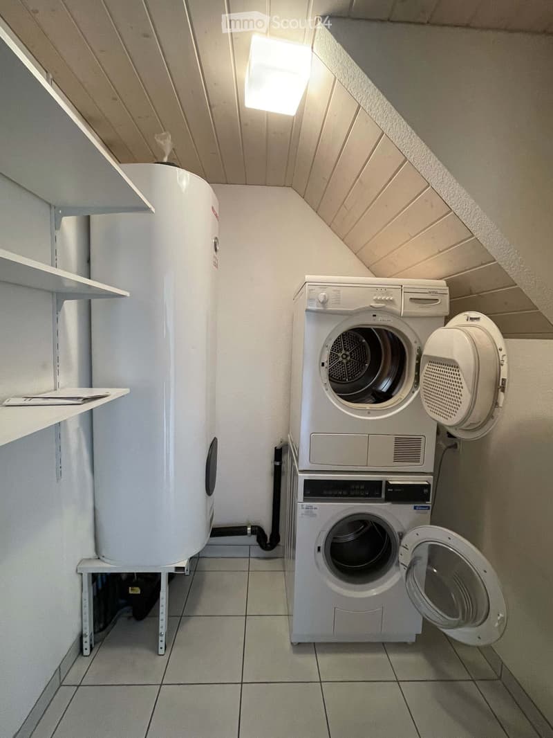 Small room with white tiled floor, white walls, ceiling with wooden panels, white shelves, white washing machine, white dryer, white water heater