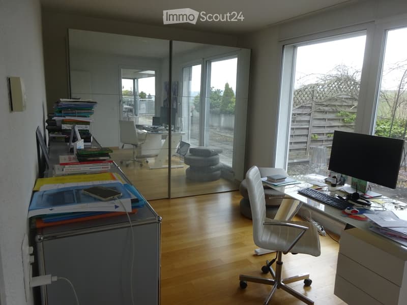 Modern office space with wooden flooring, large windows, and a mirrored wall. Contains desks, chairs, laptops, and office supplies.