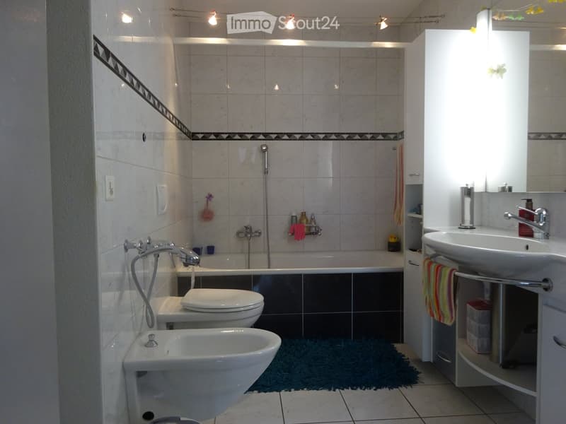 Modern bathroom with white tiles, a bathtub, toilet, sink, and a shower area. Features include a towel rack, a mirror, and a small shelf with toiletries.