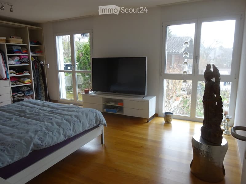Bright bedroom with wooden flooring, large windows, a double bed with blue bedding, a wardrobe, a TV on a stand, a decorative statue on a round table, and a small candle holder.