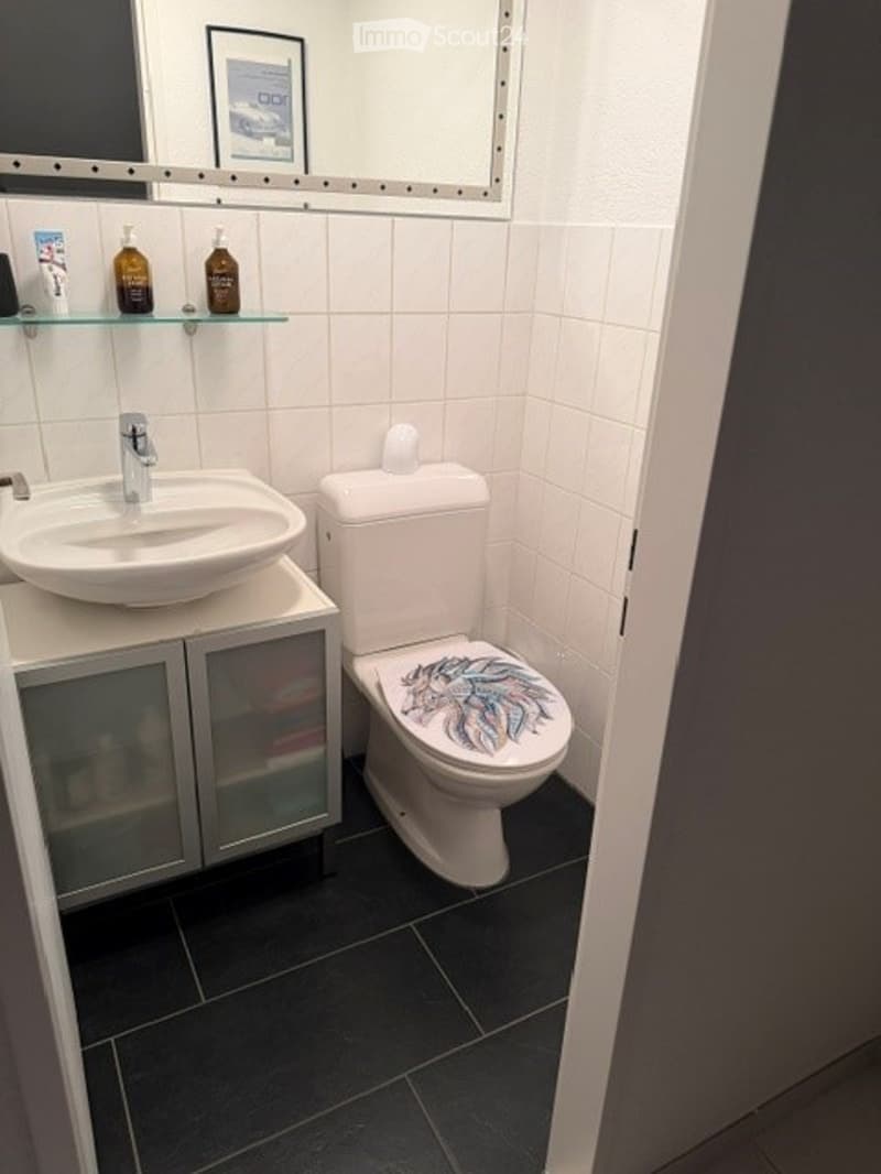 Modern bathroom with white tiles, black floor, a sink with a silver faucet, a toilet with a decorative seat cover, a mirrored cabinet, and a glass shelf holding bottles.