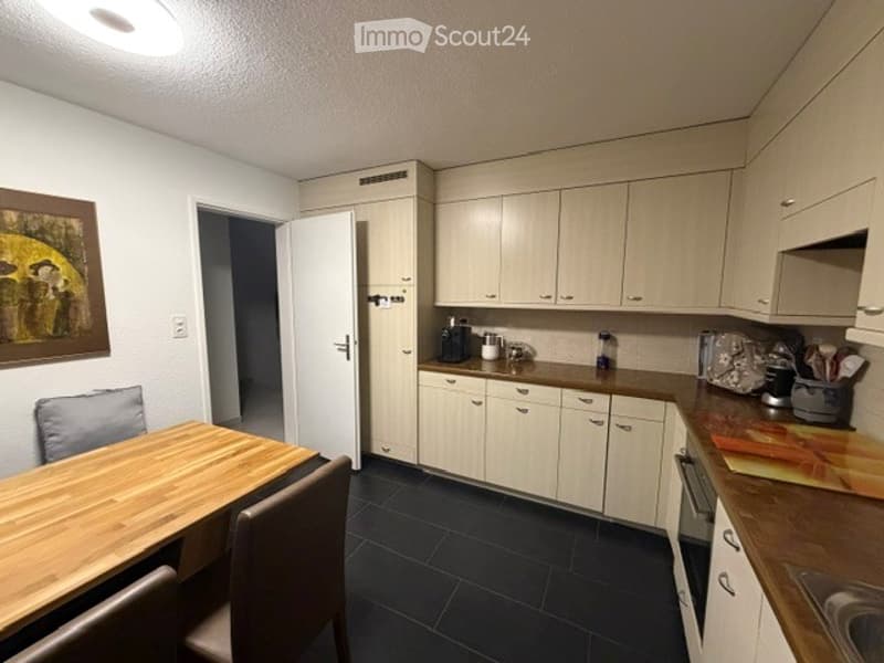 Modern kitchen with light-colored cabinets, dark tiled floor, and a small dining area with a wooden table and chairs.