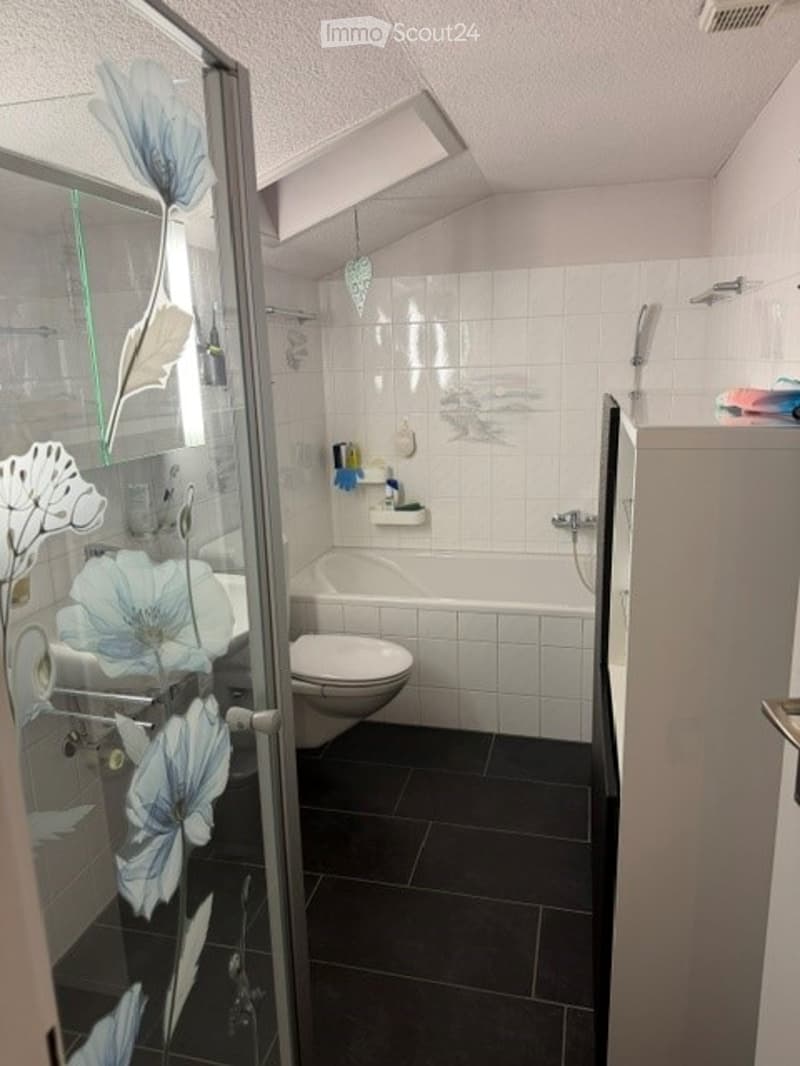 Modern bathroom with white tiled walls, black tiled floor, bathtub, toilet, glass shower enclosure with floral design, and a small shelf with toiletries.