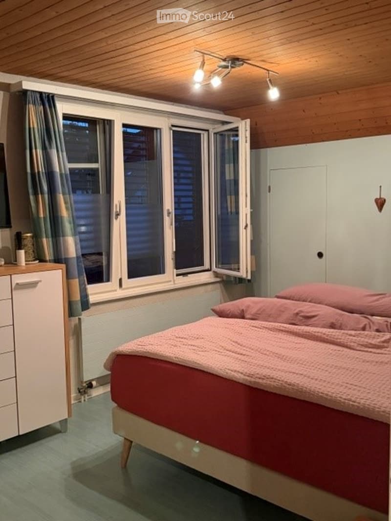 Cozy bedroom with wooden ceiling, large window with open shutters, bed with pink bedding, nightstand, and wardrobe.