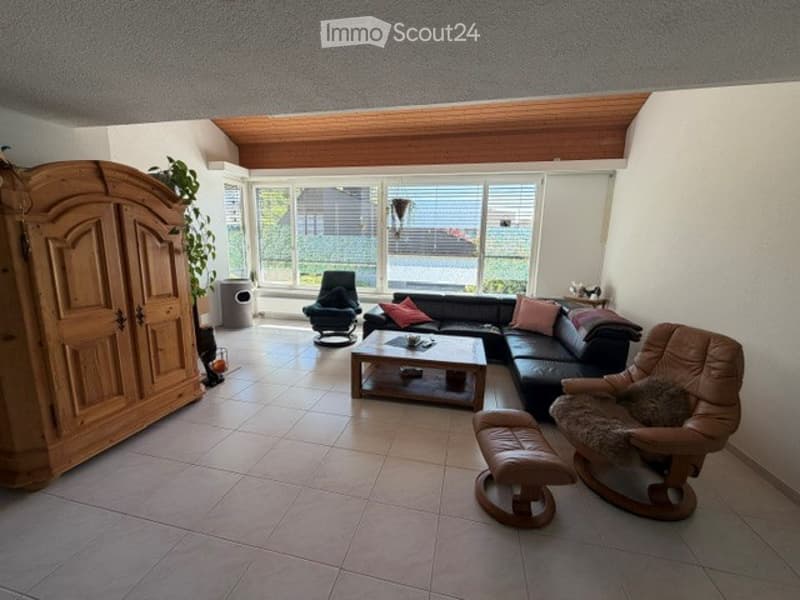 Bright living room with large windows offering a view, featuring a black leather sofa, wooden coffee table, armchair, ottoman, floor lamp, wooden cabinet, and a potted plant.