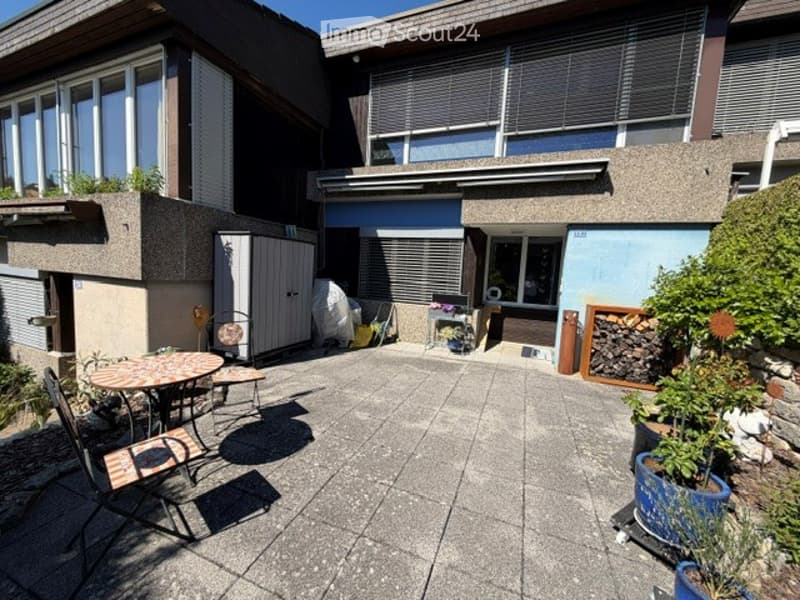 Courtyard area with patio furniture, small table, chairs, and potted plants. Adjacent to a modern house with large windows and a garage.