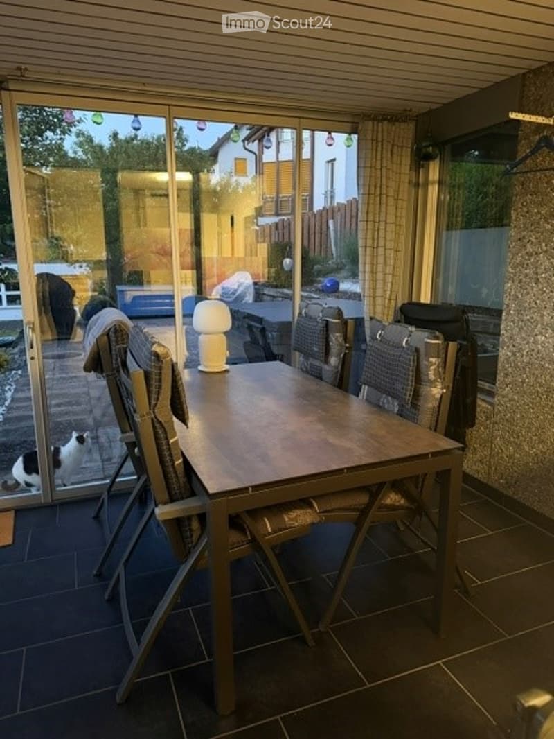 A modern dining area with a wooden table, six chairs, and a white lamp on the table. Large glass doors lead to an outdoor patio with a view of the garden.