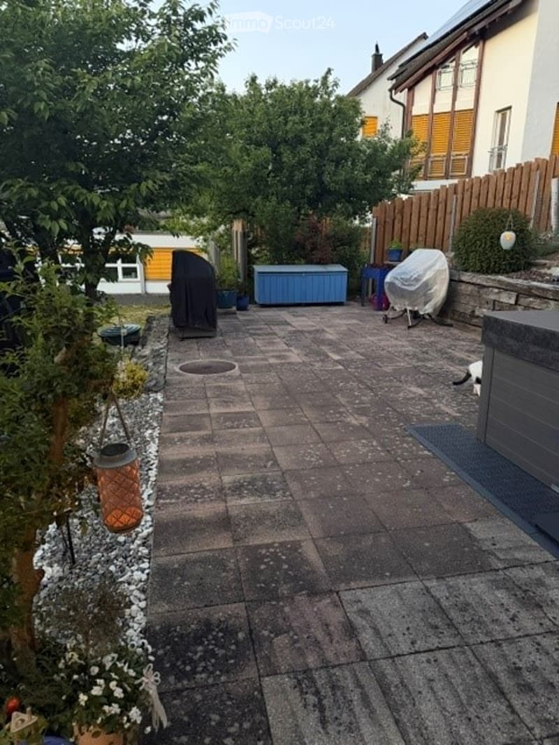 Backyard patio with stone tiles, small garden area with plants and flowers, wooden fence, and outdoor furniture including a covered grill and storage box.