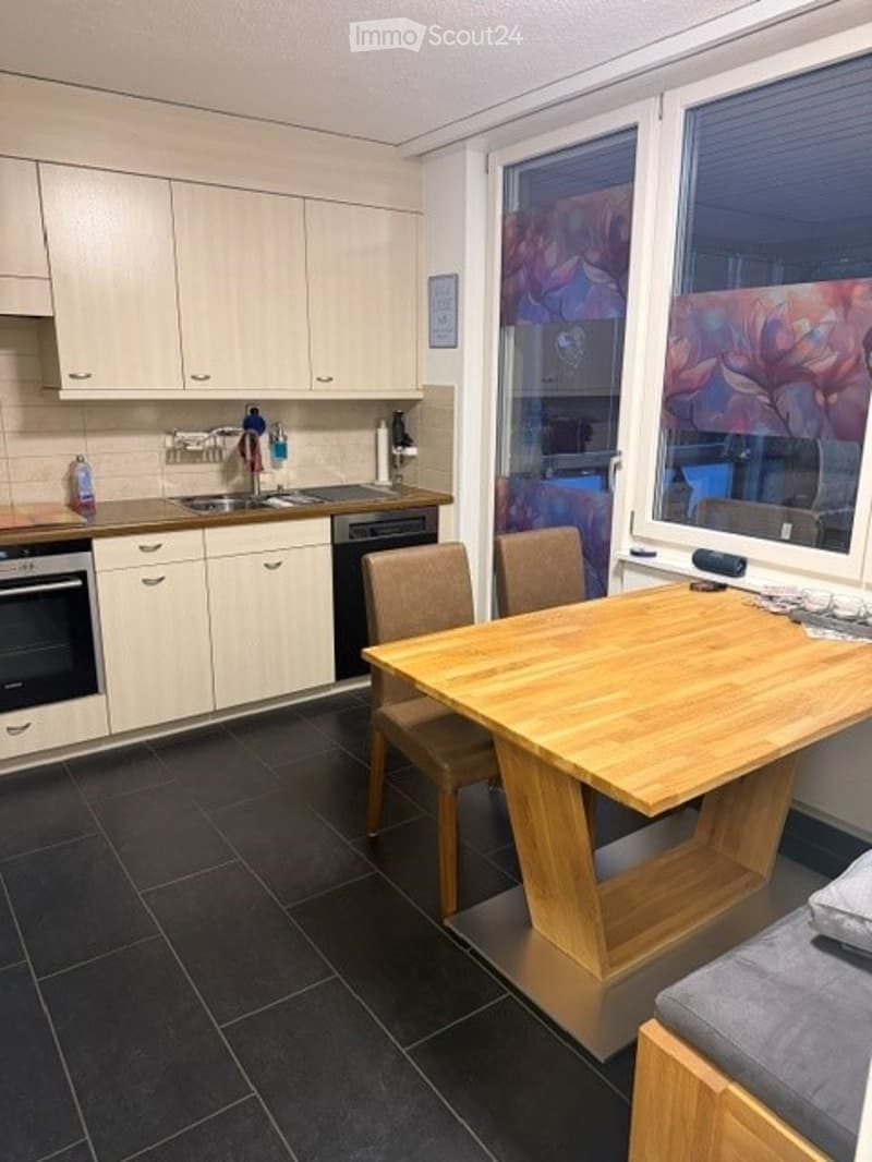 Modern kitchen with light-colored cabinets, dark tiled floor, and a wooden dining table with chairs. Features a sink, oven, and dishwasher.