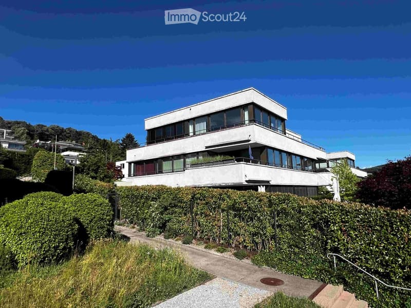 Modern two-story house with large windows and balconies, surrounded by greenery and a well-maintained hedge.