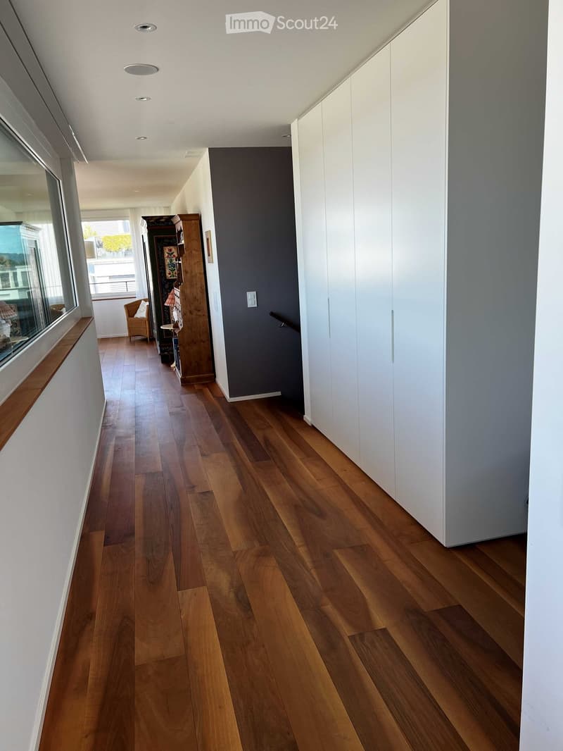 Modern hallway with wooden flooring, white walls, built-in white cabinets, and a small seating area with a chair and small table near a large window.