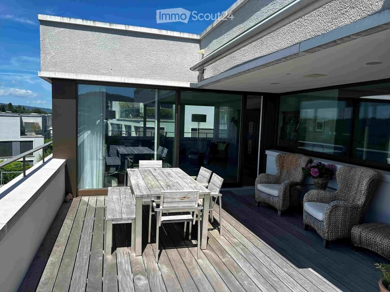 Wooden deck with a dining table, chairs, wicker chairs, and a flower pot, with large glass doors leading to the interior.