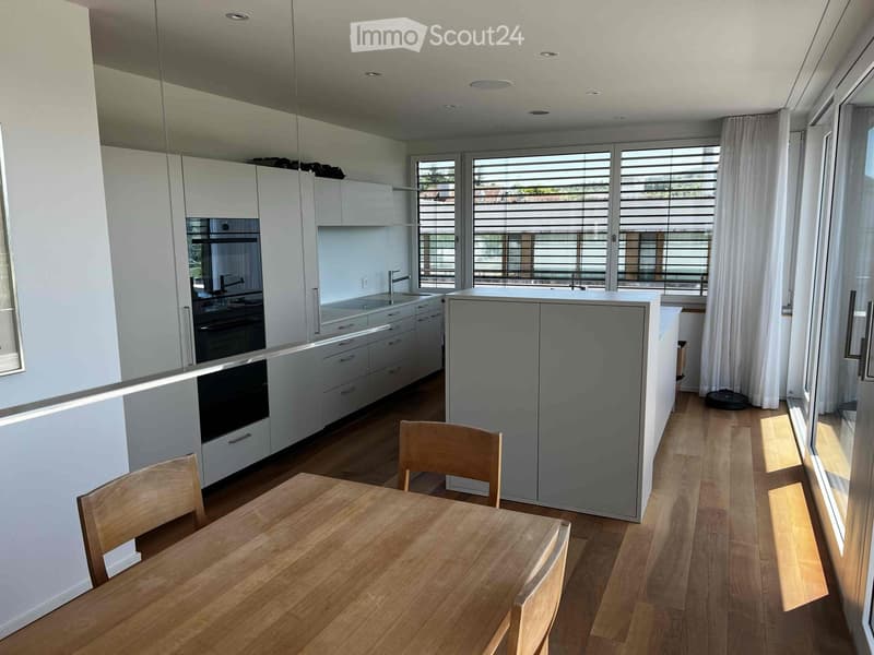 Modern kitchen with white cabinets, a wooden dining table, and chairs, large windows with blinds, and a central island.