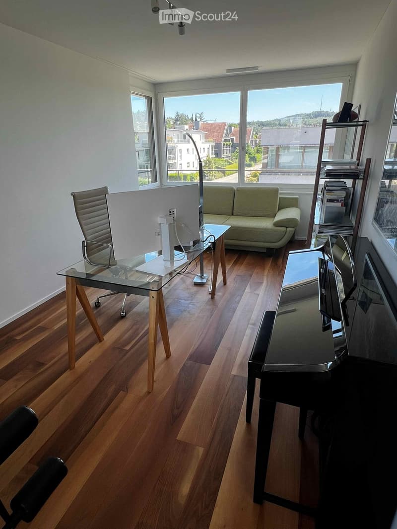 Modern, well-lit room with wooden flooring, large windows, a sofa, a glass desk, a chair, and a floor lamp.