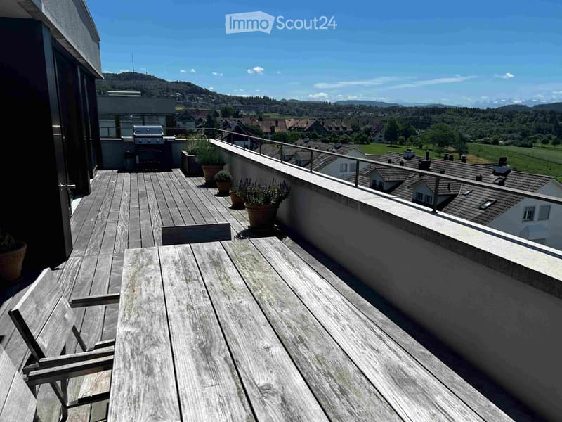 Wooden balcony with a table, chairs, potted plants, and a grill, overlooking a town and mountains.