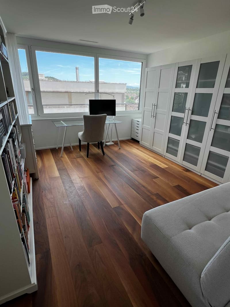 Modern living room with wooden flooring, large windows, a sofa, a chair, a glass table, a screen, and a cabinet.