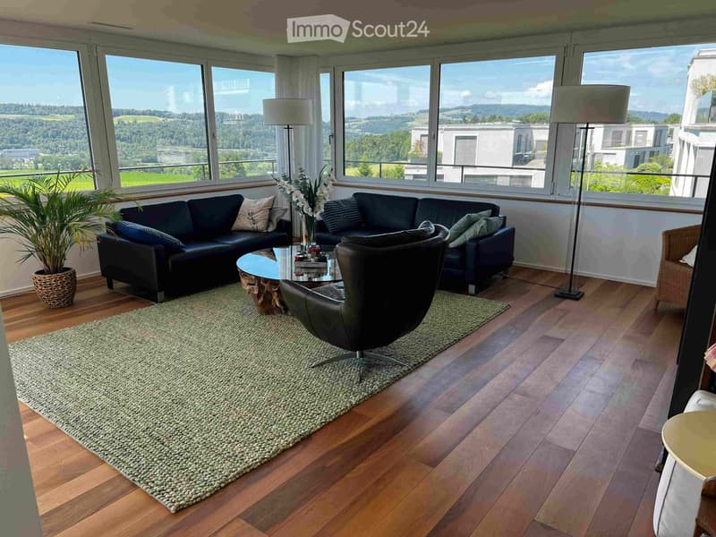 Modern living room with large windows offering a scenic view, featuring a black sectional sofa, a glass coffee table, a leather armchair, a floor lamp, a woven rug, and a potted plant.