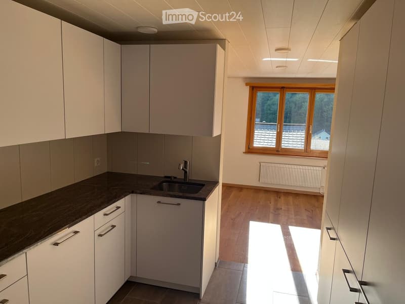 Empty kitchen with white cabinets, dark countertop, sink, and large windows letting in natural light. Wooden flooring and tiled section visible.
