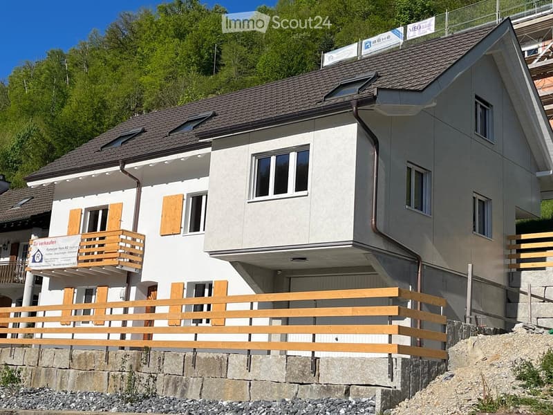 Modern two-story house with grey siding, multiple windows, wooden balconies, and a stone retaining wall.
