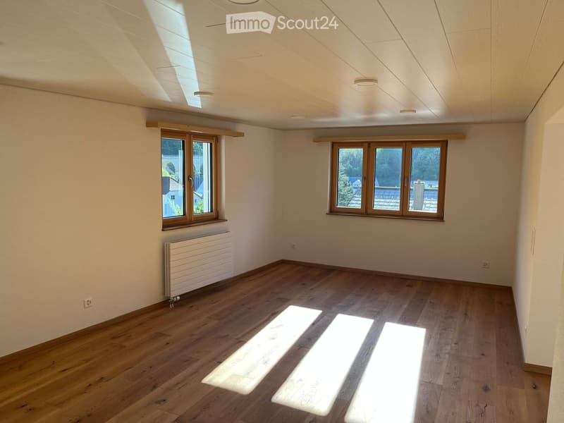Empty room with wooden flooring, white walls, and large windows allowing natural light to enter. The room features a radiator on one wall and recessed ceiling lighting.