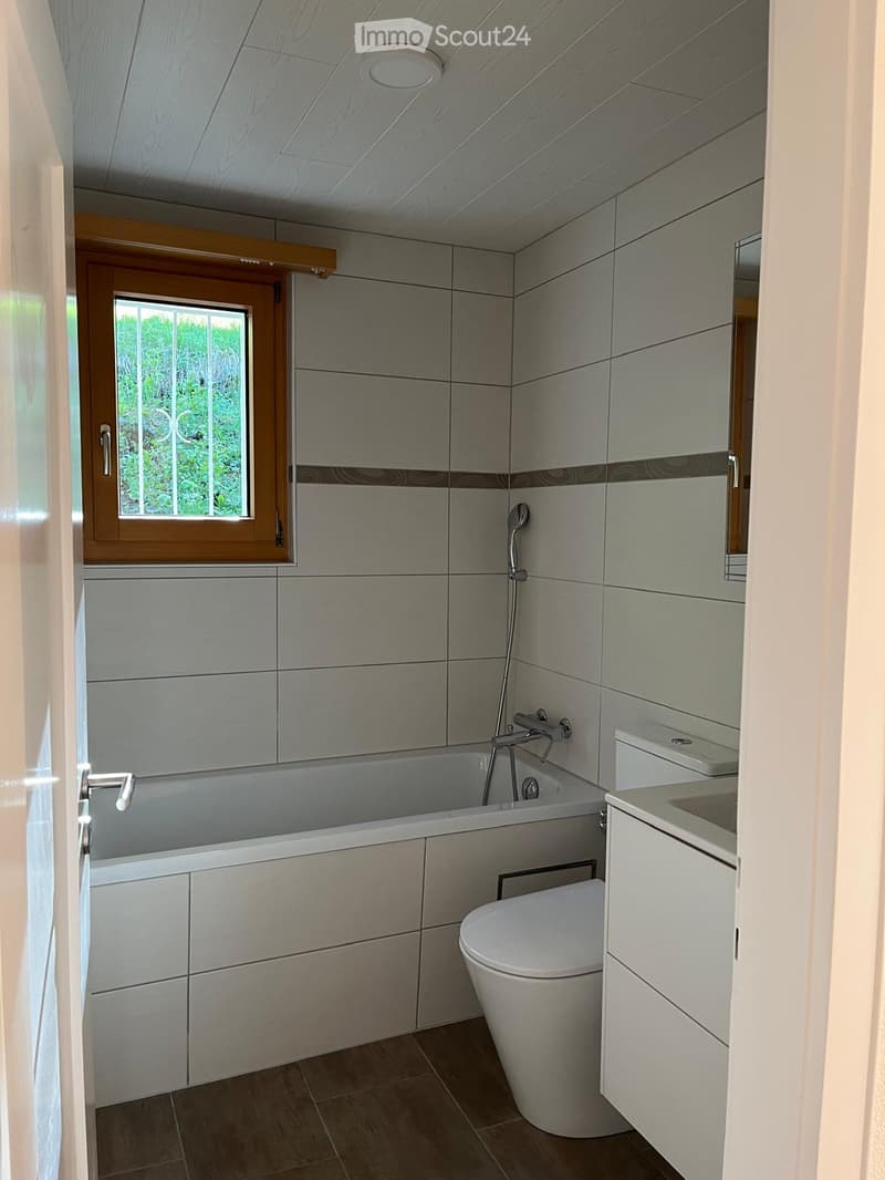 Modern bathroom with a bathtub, toilet, sink, and showerhead. Grey tiled walls and wooden ceiling.