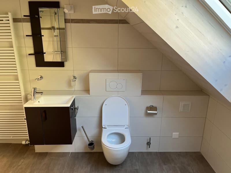 Modern bathroom with a toilet, sink, mirror, and cabinet. The room features a sloped ceiling and tiled walls.