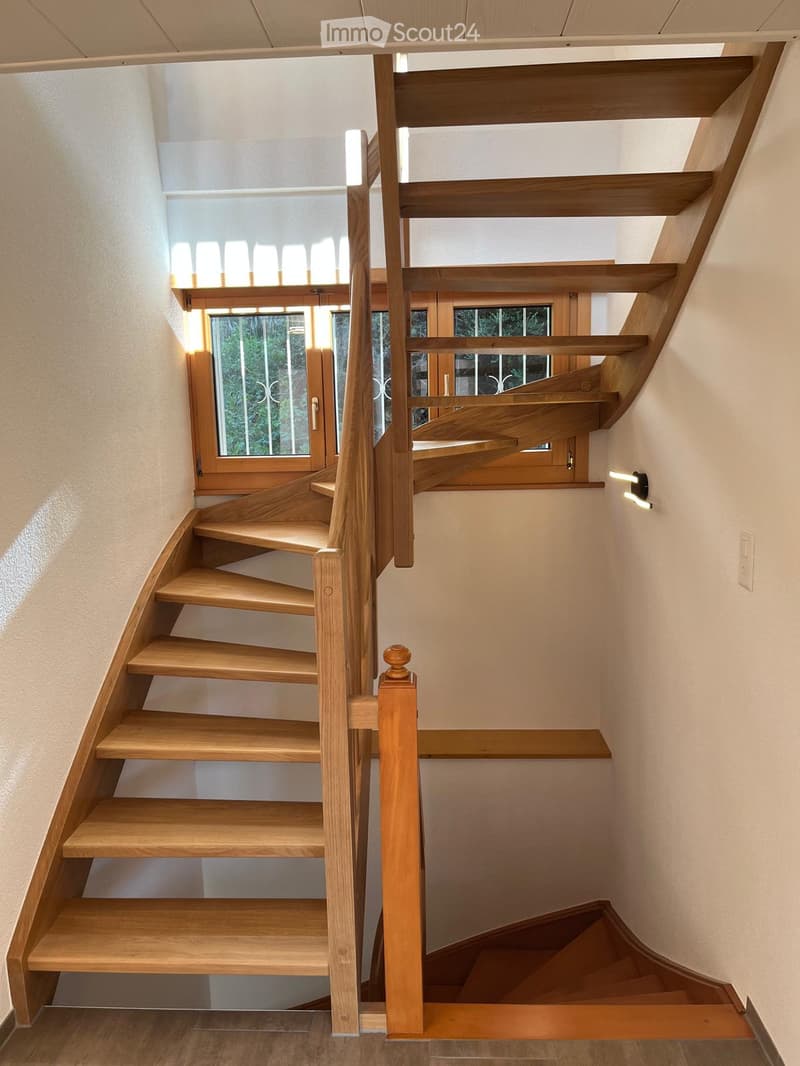 Spiral staircase with wooden steps and handrail, illuminated by natural light from a window, leading to an upper level.