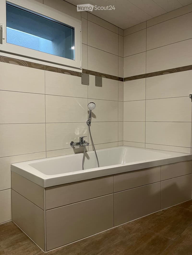 Modern bathroom with a bathtub, faucet, and showerhead. Light-colored tiles and a window provide natural light.