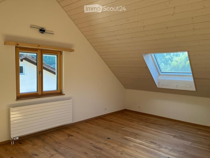 Empty attic room with wooden floors, slanted wooden ceiling, large windows, radiator, and modern lighting fixture.