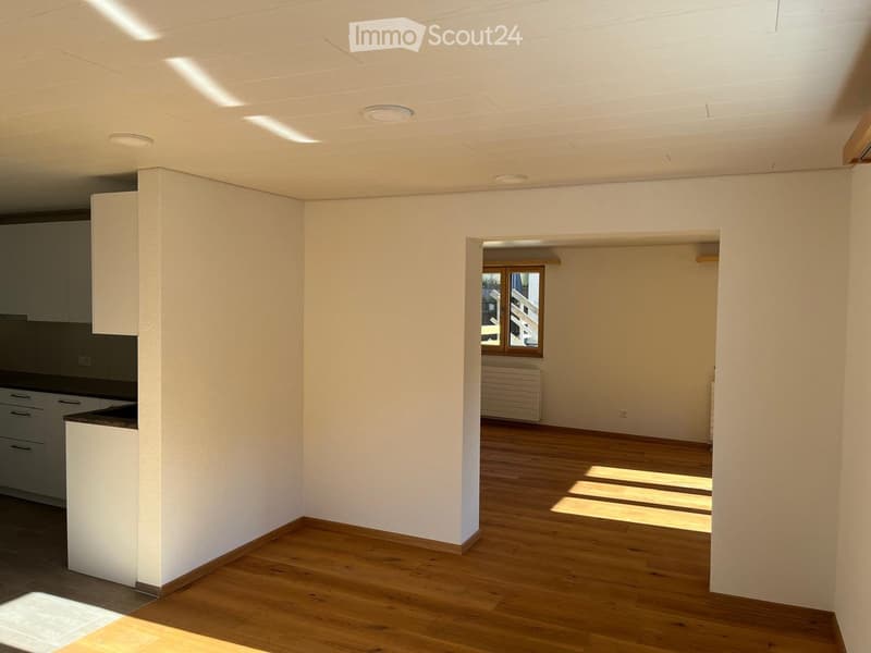 Empty room with wooden flooring, white walls, and a small kitchen area visible in the background. The space is well-lit with natural light coming through a window.