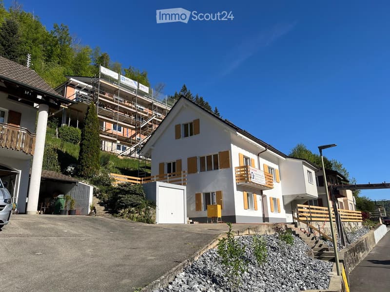 Two-story residential building with white walls, yellow shutters, and balconies. The house has a garage and is surrounded by greenery and a paved driveway.