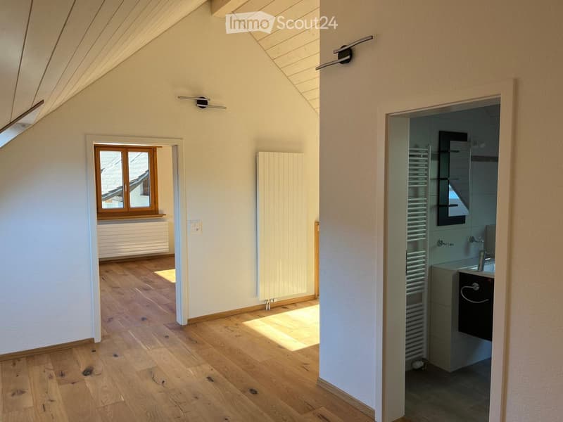 Bright, empty attic room with wooden flooring, white walls, and a skylight window. A small bathroom is visible through an open doorway.