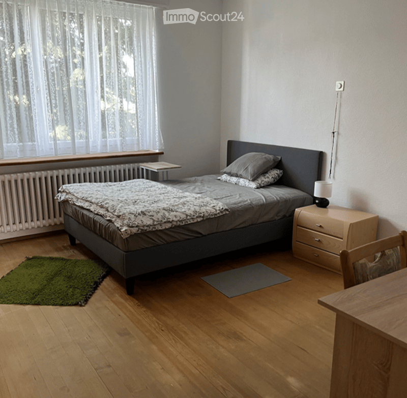 Cozy bedroom with wooden flooring, a grey bed with pillows and a blanket, a nightstand with a lamp, a small table, and a green rug.