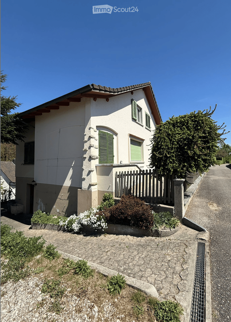 Two-story house with white exterior, green shutters, and a small garden area with shrubs and flowers.