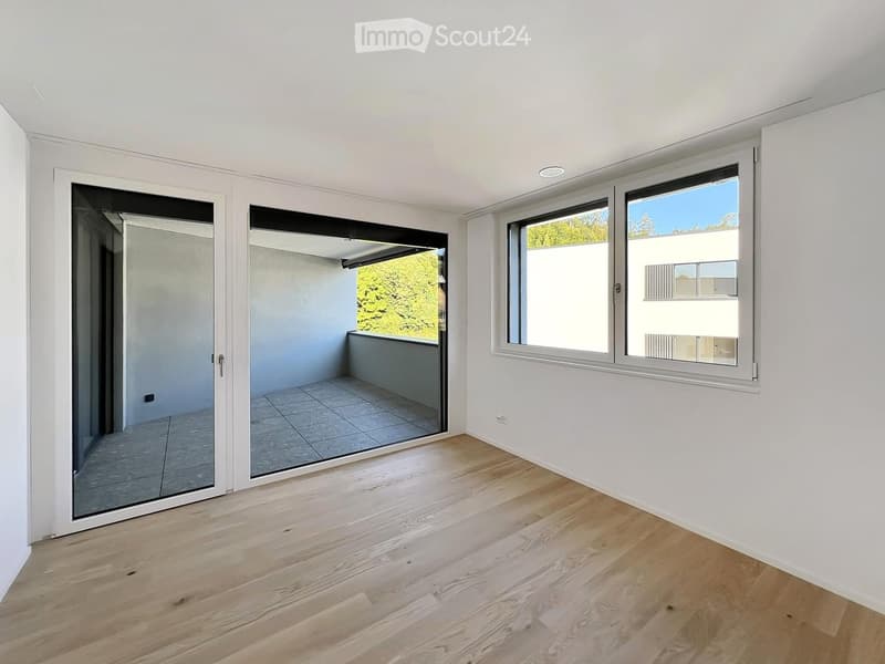 Empty room with light wooden flooring, white walls, large windows, and sliding glass doors leading to a balcony.
