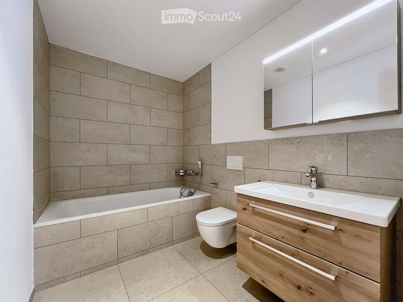 Modern bathroom with beige tiled walls and floor, a bathtub, toilet, sink with wooden cabinet, and a large mirror with LED lighting.