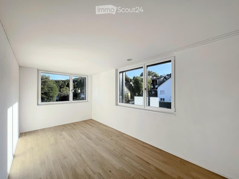 Empty room with light wooden flooring and white walls, featuring two large windows with white frames that offer views of greenery and neighboring houses.