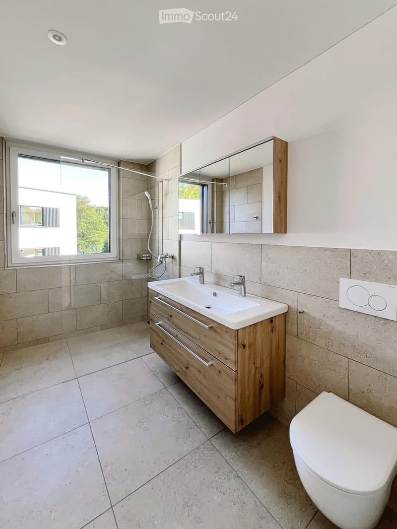 Modern bathroom with a white sink, wooden cabinet, mirror, toilet, and shower area with a window providing natural light.