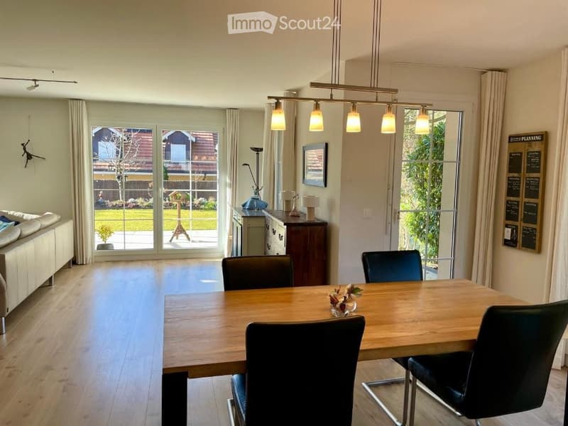 Bright, open-plan living and dining area with wooden flooring, large windows, and sliding glass doors leading to a garden. Features a cream sofa, a wooden dining table with four black chairs, a sideboard, and a hanging light fixture.