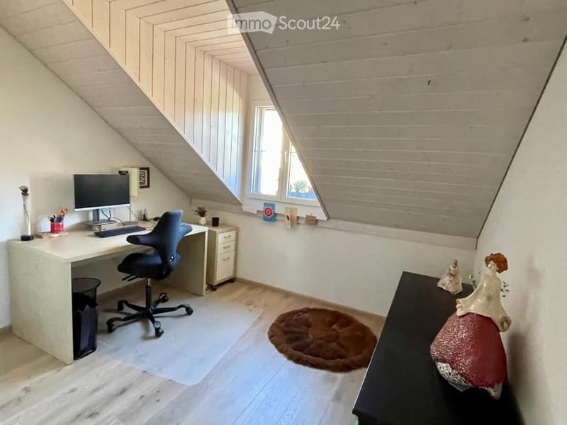 A cozy attic room with slanted wooden ceilings, a large window, a desk with a computer, a chair, a small cabinet, a round rug, and decorative items on a table.
