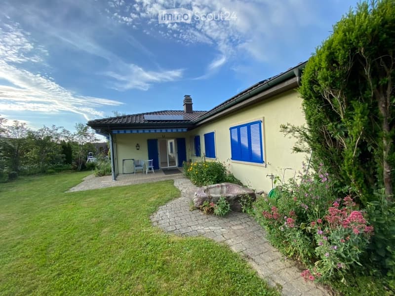 Single-story house with light yellow walls, blue shutters, and a covered entrance area. The garden features a well-maintained lawn, a stone pathway, and colorful flowering plants.