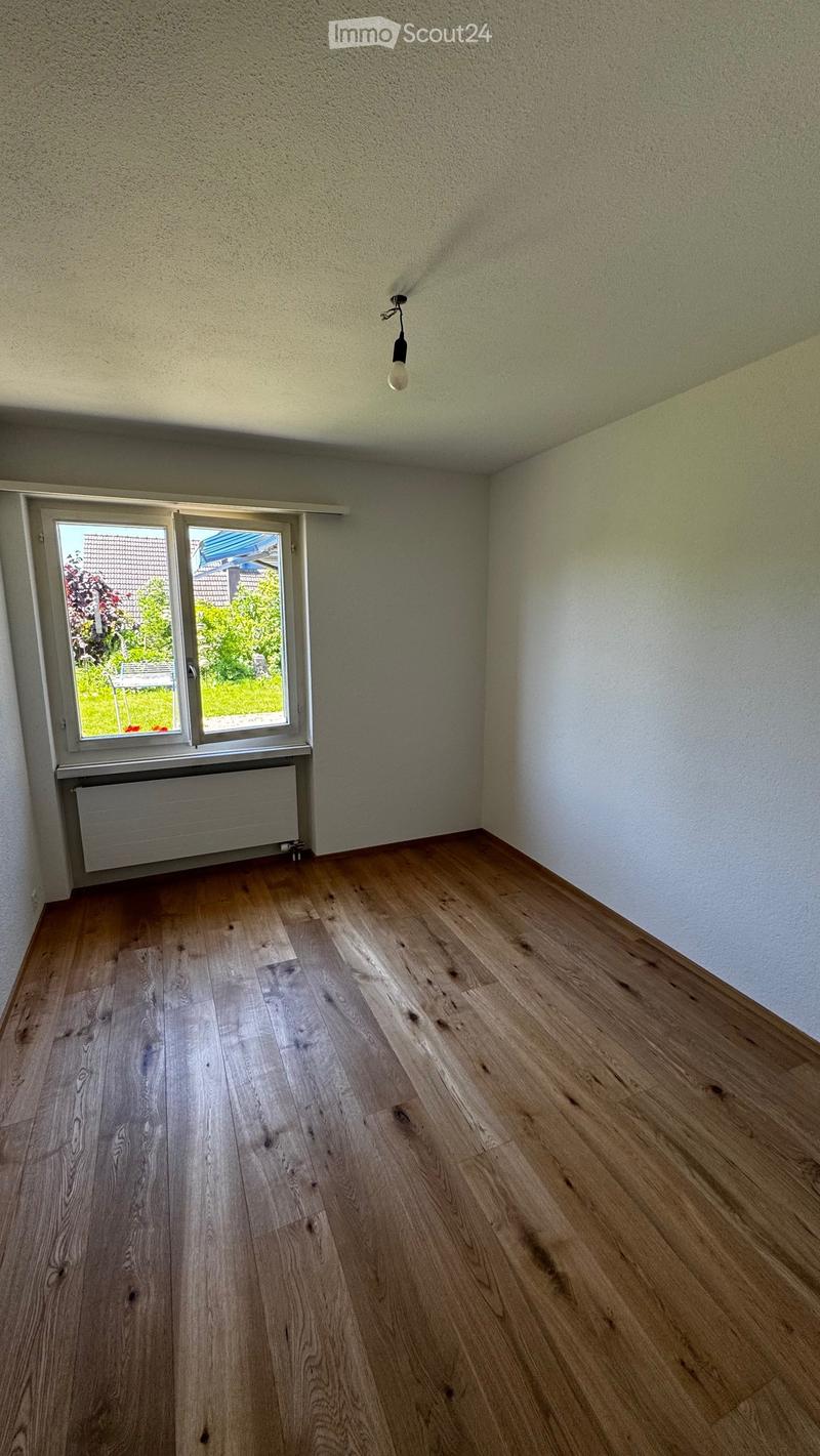 Empty room with wooden flooring, white walls, and a single window with a radiator below it. The room has a simple, clean design with no furniture or decorations.