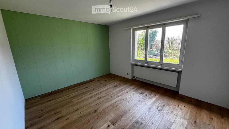 Empty room with wooden flooring, light grey walls, and a large window with a view of greenery outside. A radiator is visible beneath the window.