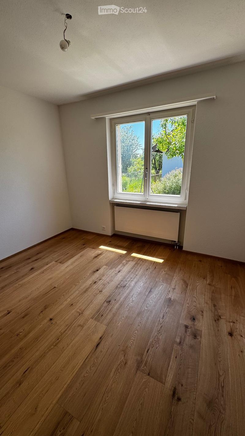 Empty room with wooden flooring, white walls, a window with white frame, radiator, and a light fixture on the wall.