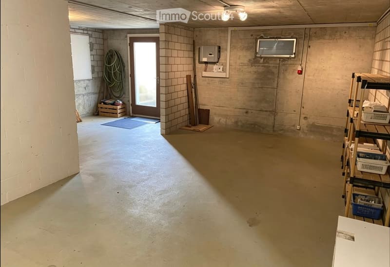 Empty basement or utility room with concrete walls and floor, a door leading outside, and minimal storage items like a shelf and a few boxes.