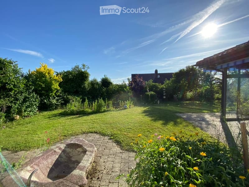 Well-maintained garden with a lawn, flower beds, a stone fire pit, and a paved pathway leading to a house with a covered patio area.