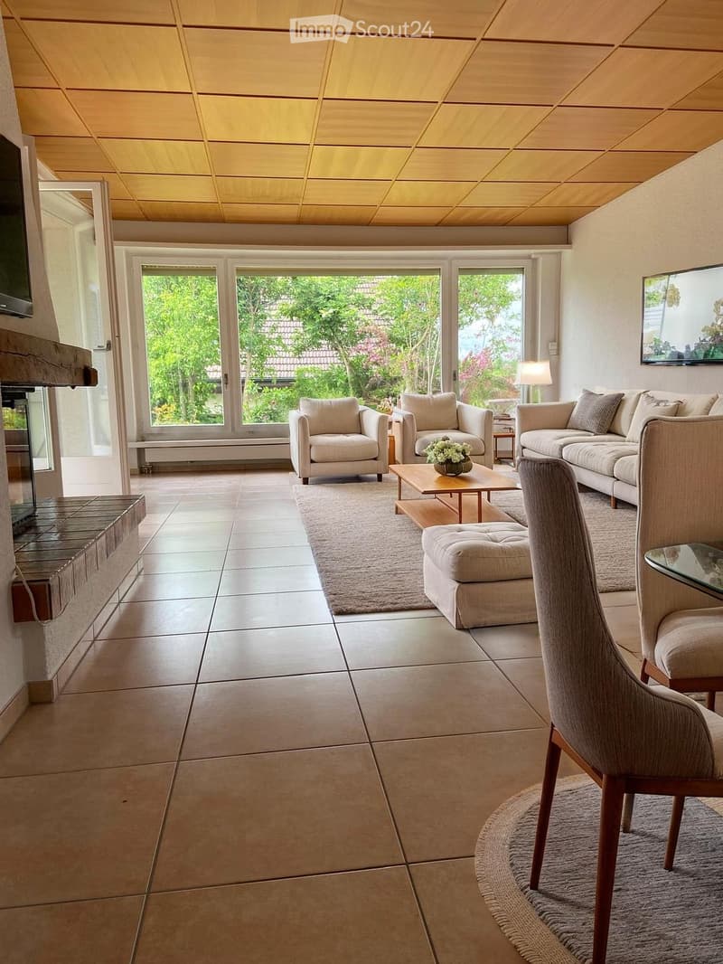 Bright living room with large windows and a view of greenery, featuring a beige sofa, armchairs, coffee table, ottoman, and a fireplace.