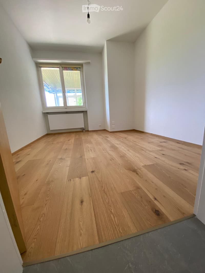 Empty room with light wooden flooring, white walls, and a window with blinds. The room has a radiator below the window and a visible electrical outlet on the wall.