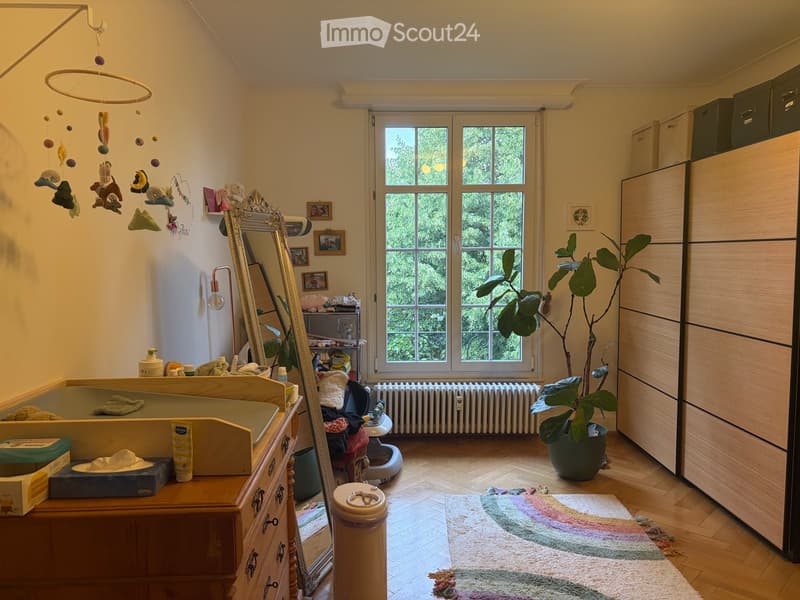 A well-lit children's room with a wooden cradle, a large mirror, a potted plant, and a colorful rug on the floor. The room features a wooden cabinet, a toy mobile hanging from the ceiling, and several framed pictures on the wall.