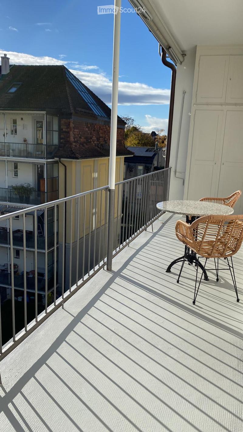 Balcony with a round table and two wicker chairs, metal railing, and a view of neighboring buildings.